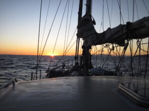 View from a sailboat at sea during sunset, with the boat's deck and rigging in the foreground, and the sun setting on the horizon over the ocean. Antarctic landscape.