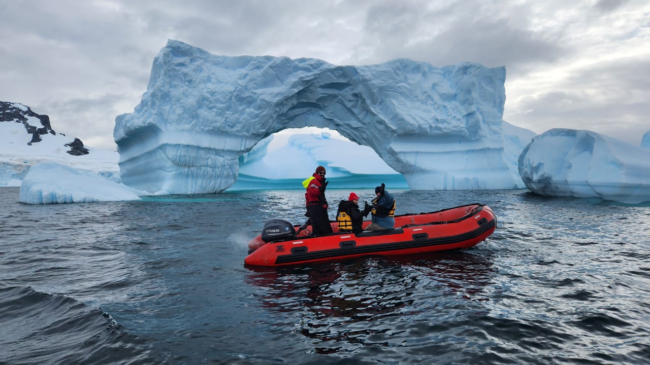 Group of people in a red inflatable boat near a large iceberg with an arch in Antarctica, with more icebergs and a cloudy sky in the background.