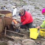Two individuals working on an archaeological excavation site with rusted metal containers, penguins on rocky terrain in the background, in South Georgia