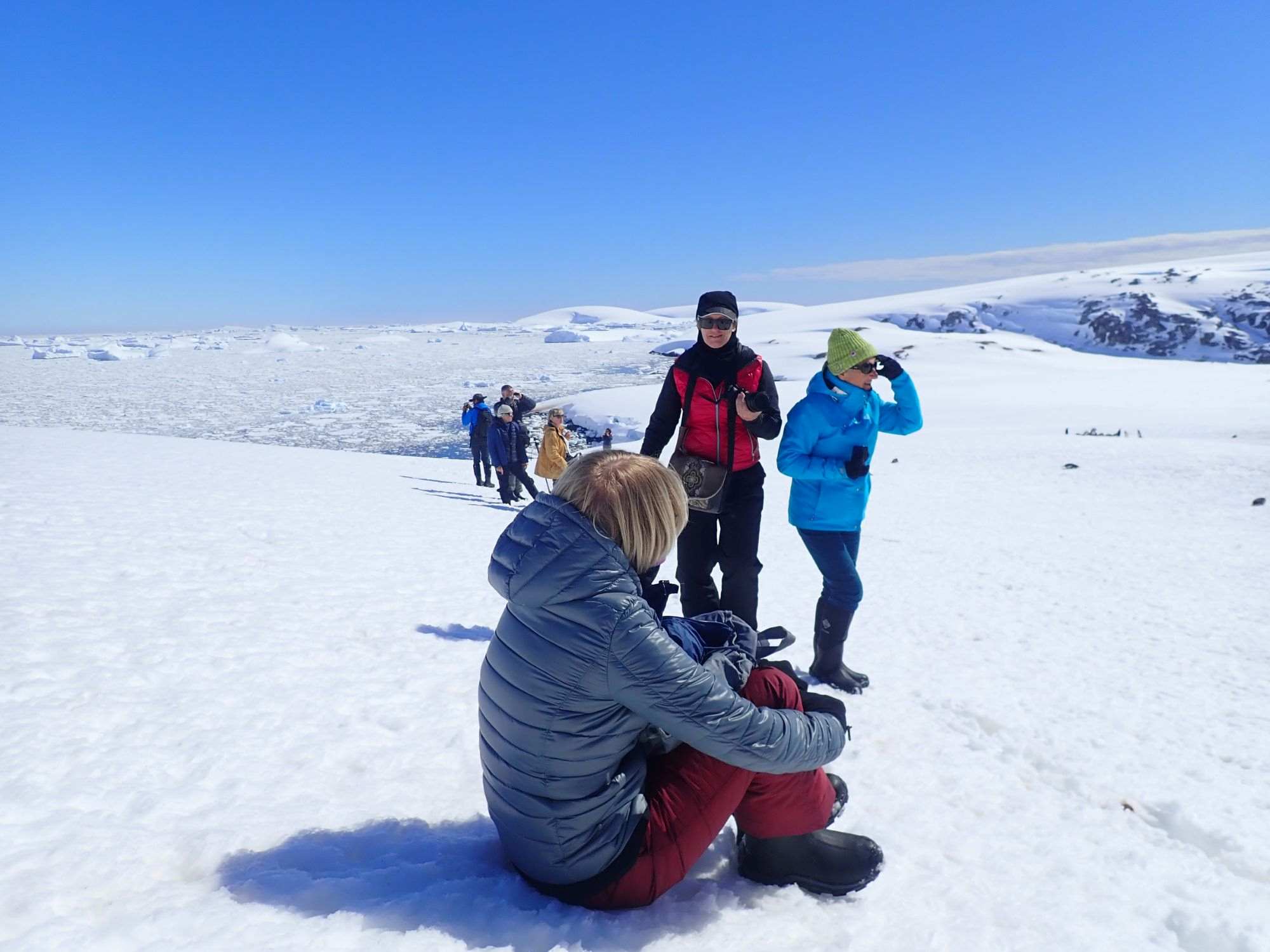Snowy Antarctic landscape with people in winter clothing walking and kneeling on the snow, distant hills in background.