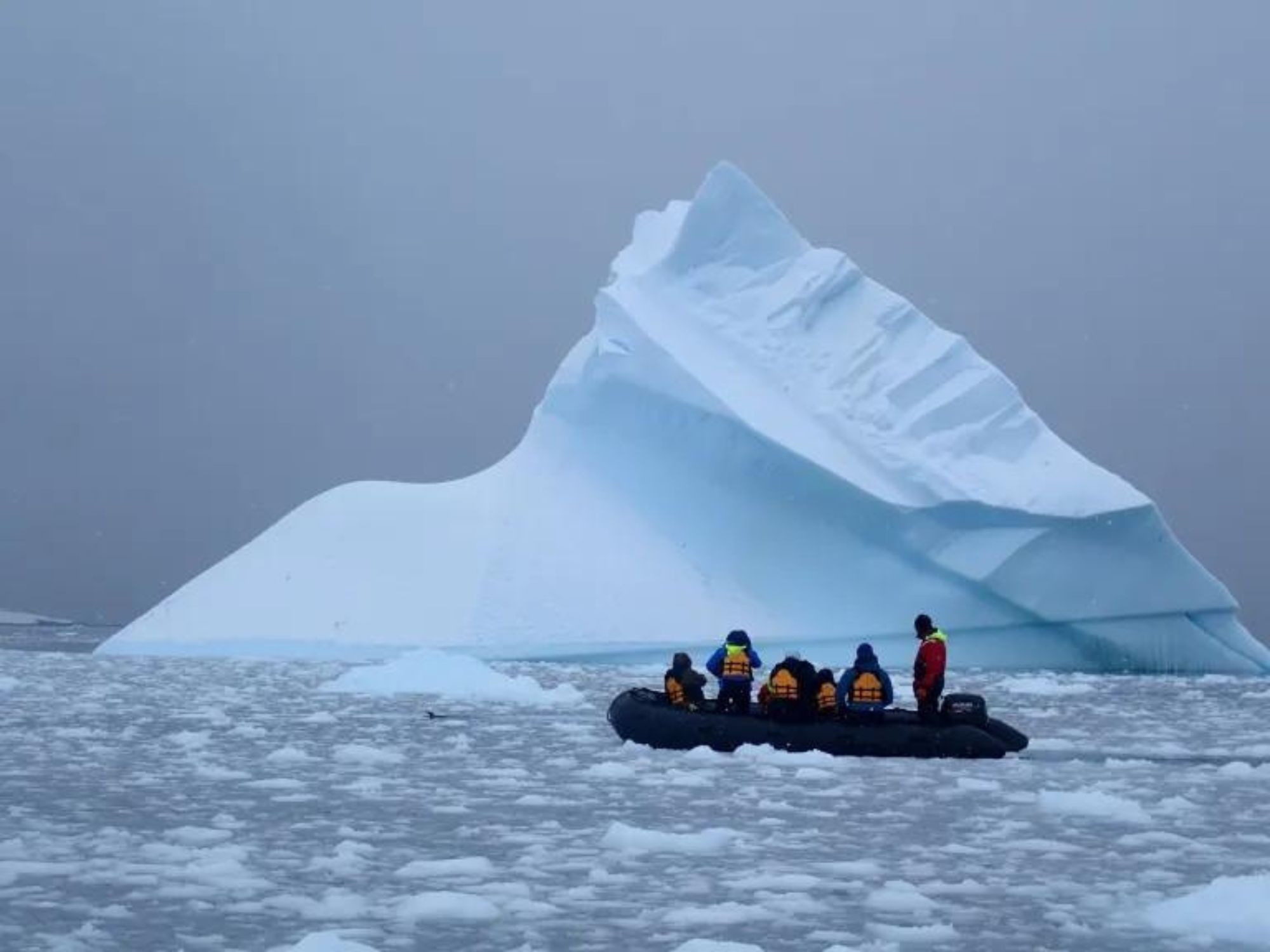 Small inflatable boat with people in life jackets navigating through icy waters, with a large iceberg in the background, Antarctica.